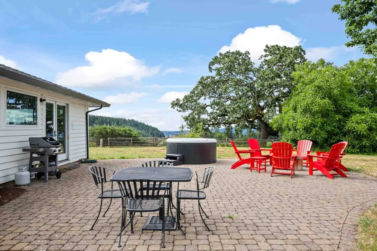 This exterior image captures a spacious patio area featuring a brand new hot tub, red Adirondack chairs, a sleek black outdoor dining set, and a large tree providing shade. The scene overlooks rolling hills and vineyard views, creating an inviting atmosphere for relaxation and social gatherings.
