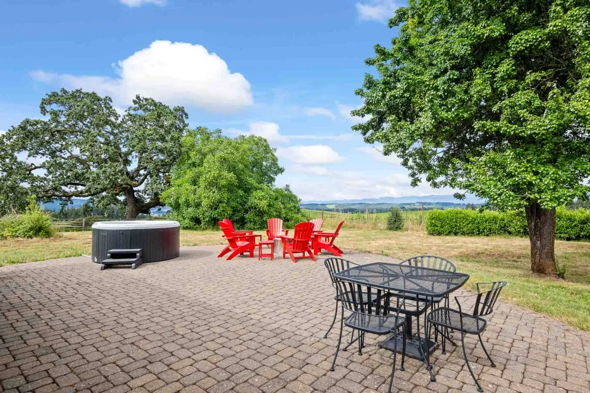 An exterior view featuring red Adirondack chairs, an outdoor dining table, a hot tub, large shade trees, and distant hills. The setup is ideal for group relaxation, outdoor meals, and soaking in scenic views, with various seating options for both adults and children.