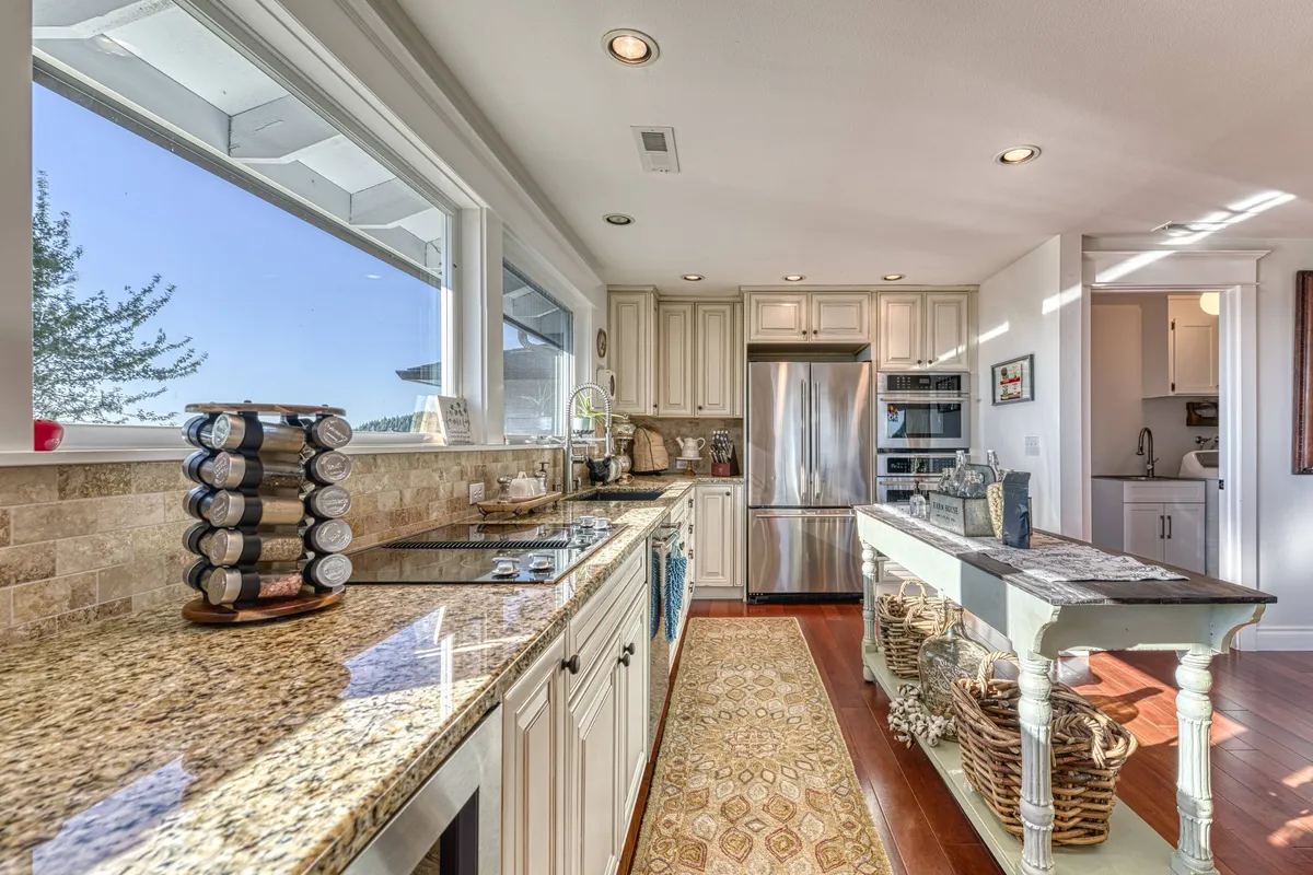 A bright, inviting kitchen with granite countertops, stainless steel appliances, a spacious island with wicker baskets underneath, and large windows bringing in abundant natural light. The setup is ready for both cooking and casual entertaining.