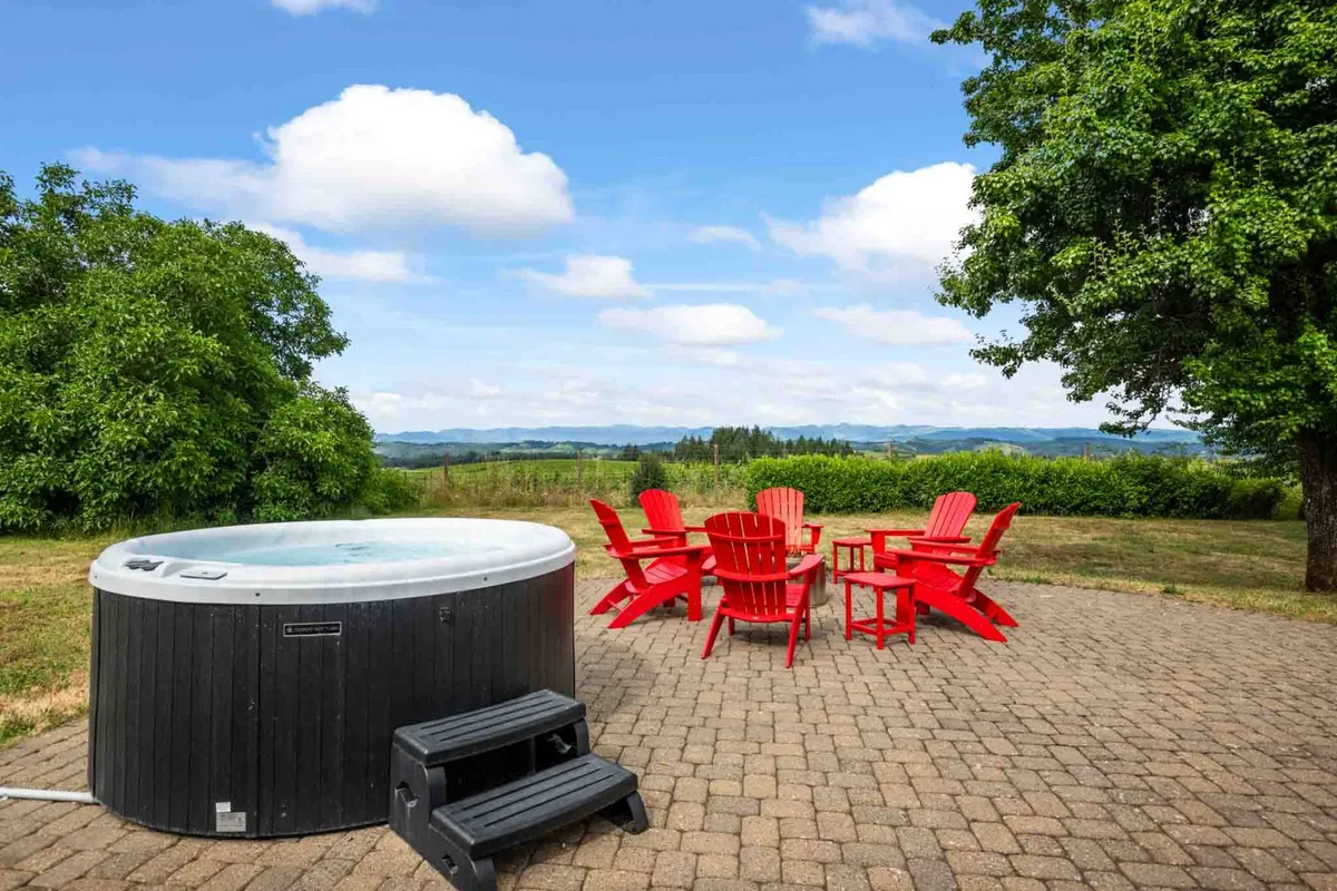 The photo highlights a private hot tub surrounded by red Adirondack chairs, all set against a landscape view. The setting radiates relaxation and social enjoyment, hinting at evening gatherings and sunset toasts after a day of wine touring.