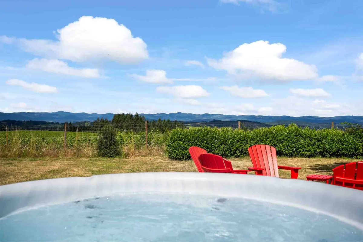 An inviting scene featuring a bubbling hot tub framed by red Adirondack chairs, overlooking rolling hills and distant mountains. The setting is surrounded by greenery and offers breathtaking panoramic views, hinting at both relaxation and awe-inspiring natural beauty.
