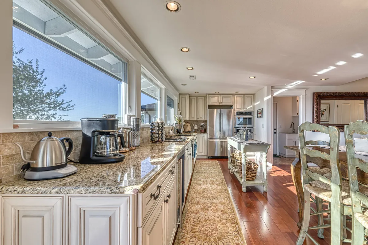 A warm, welcoming dining area adjacent to the kitchen, featuring a granite countertop, stainless steel appliances, large windows, and a wooden dining table. Abundant natural light and an open layout suggest easy flow between living, dining, and kitchen spaces.