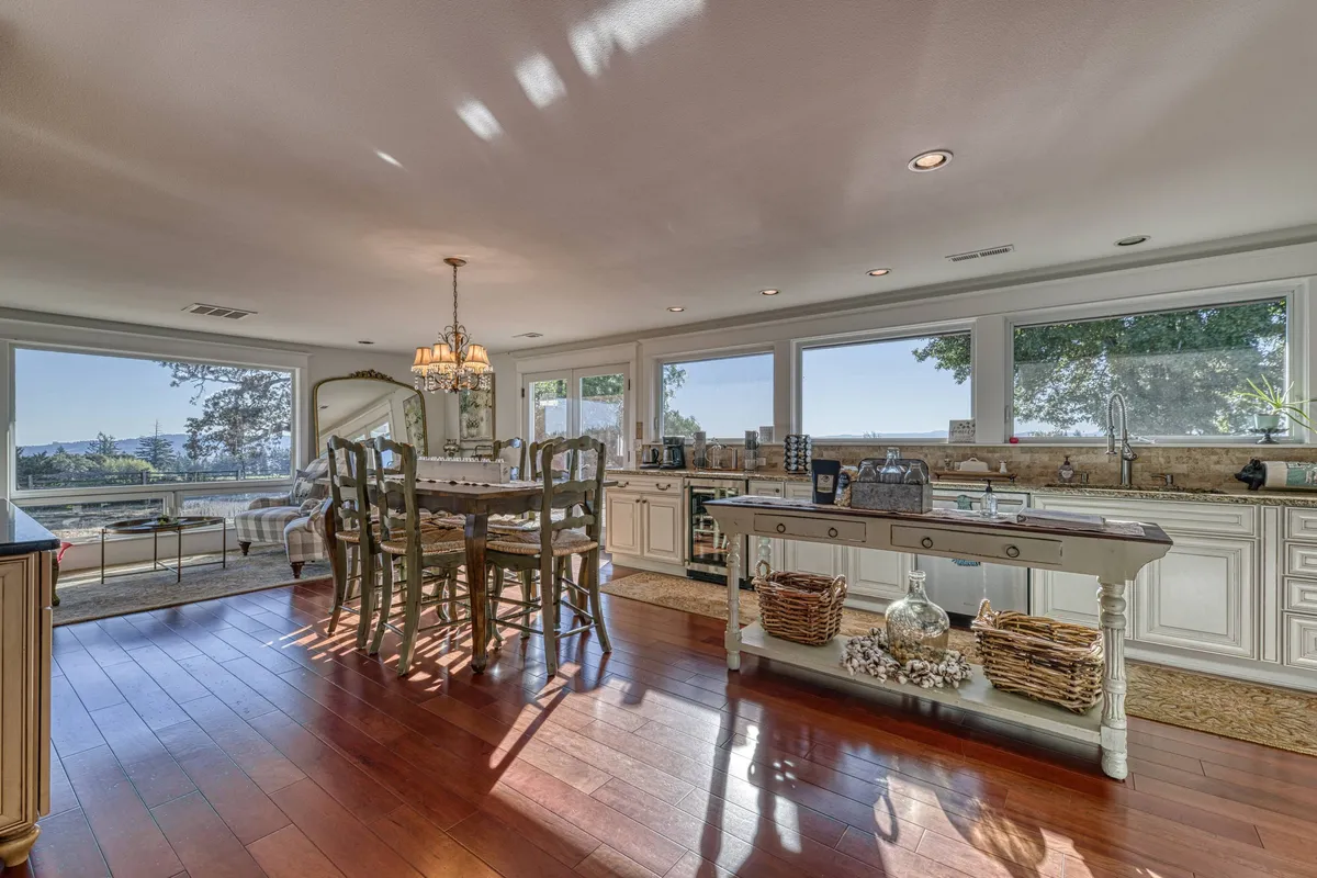 The image features a warm, elegant dining area with a stylish table set beneath a sparkling chandelier, a modern kitchen island ready for gatherings, and expansive windows revealing beautiful vineyard and hillside views bathed in sunset light.