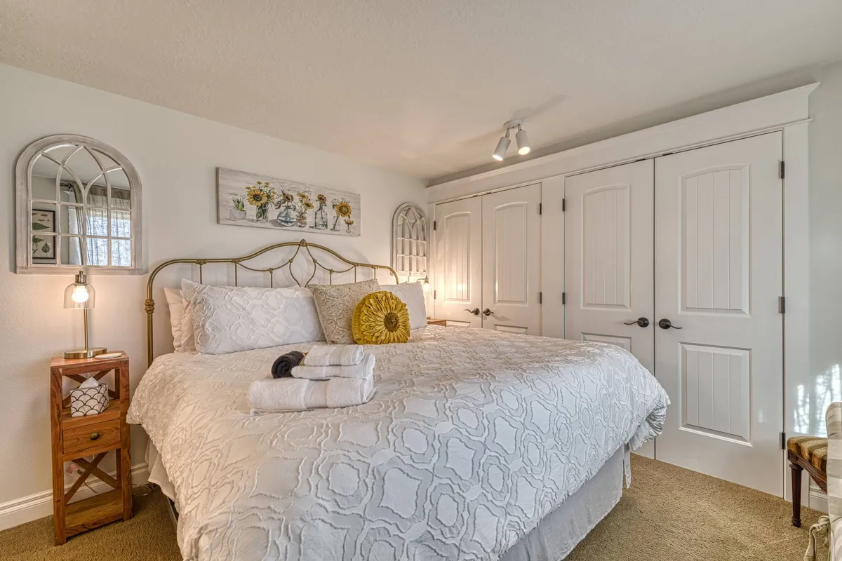 A bright and welcoming bedroom featuring a neatly made bed with decorative pillows, a nightstand with a lamp, wall mirrors, artwork, and closet doors, all within a calm, light-filled setting.