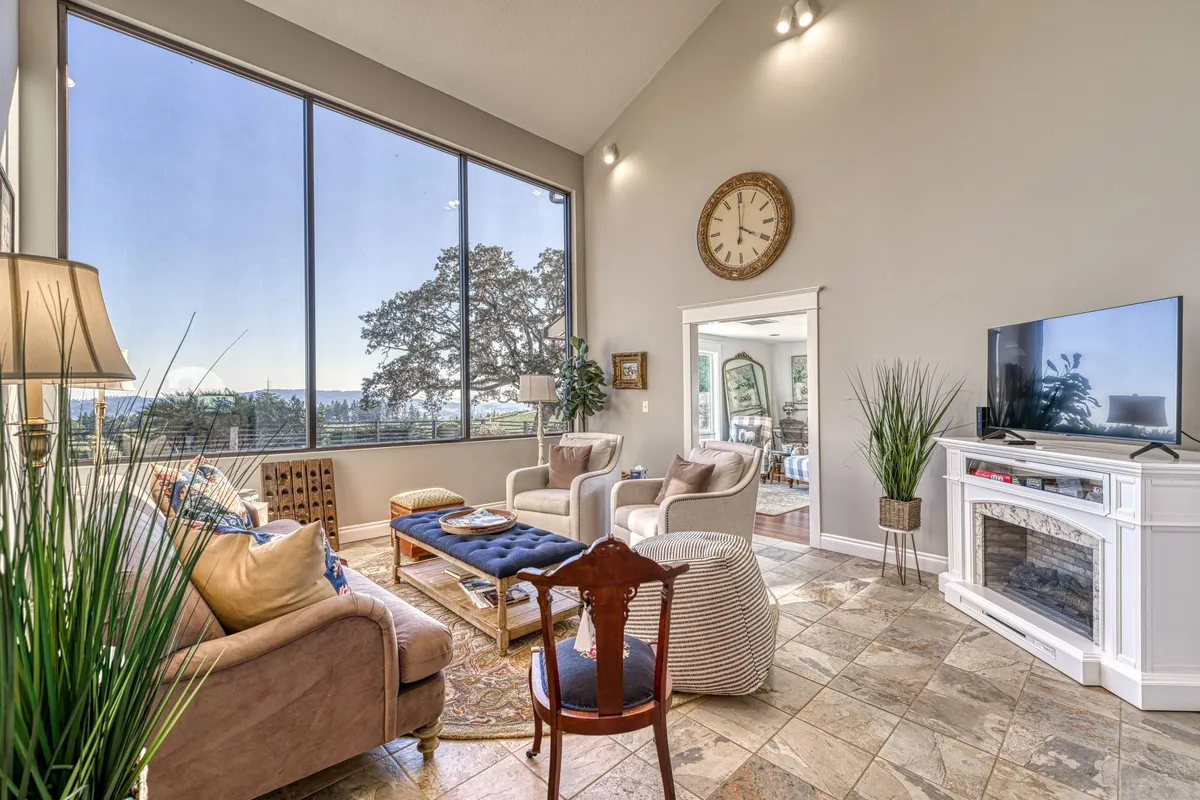 A spacious living room bathed in natural light, featuring large windows that reveal sweeping vineyard views. The room is furnished with a blend of plush, new and vintage seating, a decorative clock, a cozy electric fireplace, and a smart TV, all arranged to create an inviting atmosphere for relaxation and connection.