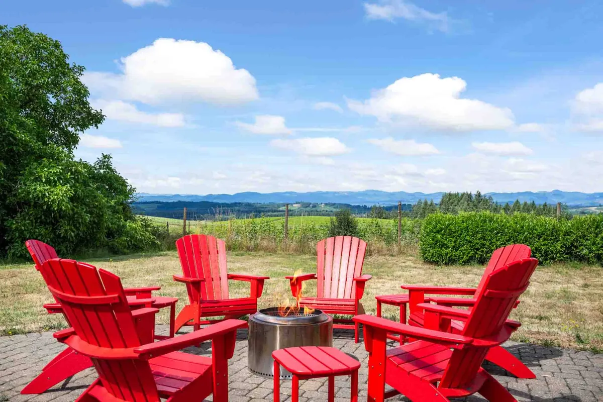 A cozy circle of red Adirondack chairs surrounds a fire pit, set against a scenic landscape with open skies and lush greenery. The arrangement suggests evenings spent storytelling and stargazing in the backyard.