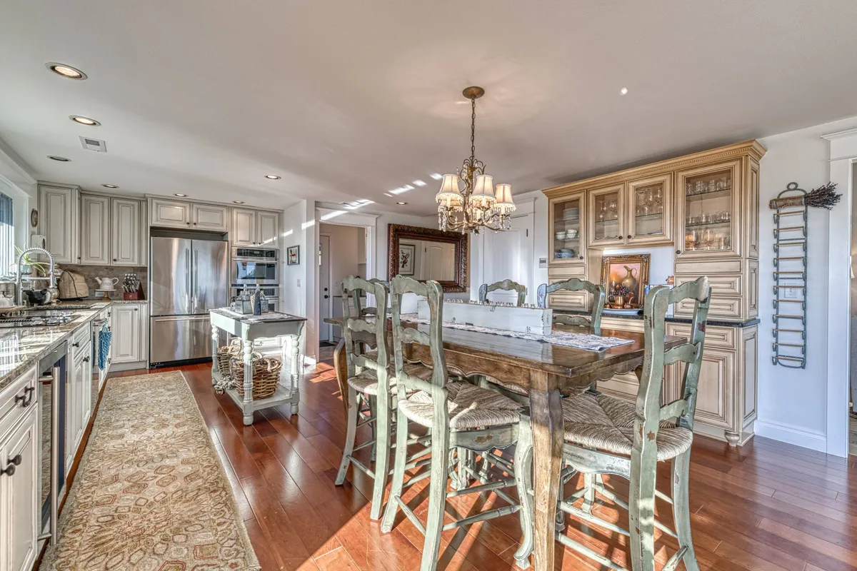 A spacious open-concept kitchen and dining area featuring a large wooden dining table under a chandelier, a modern kitchen island, stainless steel appliances, and glass-front cabinetry. The space is warmly lit and set up for both meal prep and elegant dining experiences.
